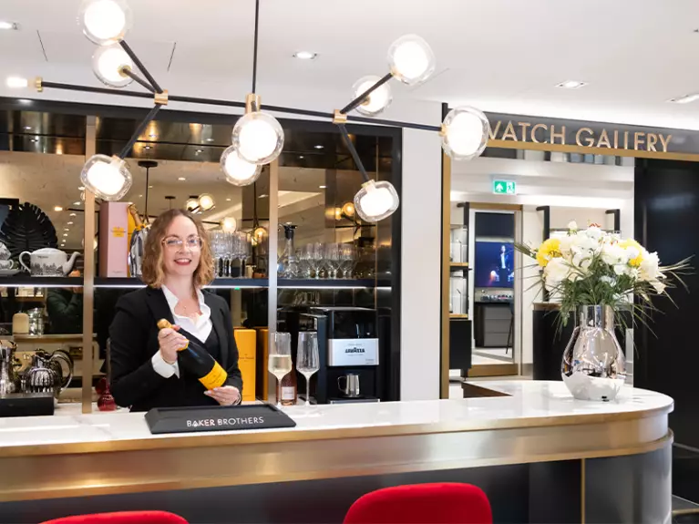 Female Baker Brothers team member holding Champagne behind the bar in front fo the watch gallery